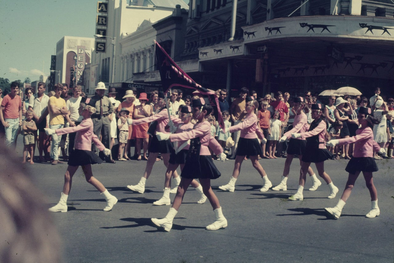 A group of eight individuals marching in a parade at The Warana Festival in the 60's. They wear matching pink jackets, black skirts, white dress boots and black and pink performance hats. A large pink and black unreadable flag is displayed. 