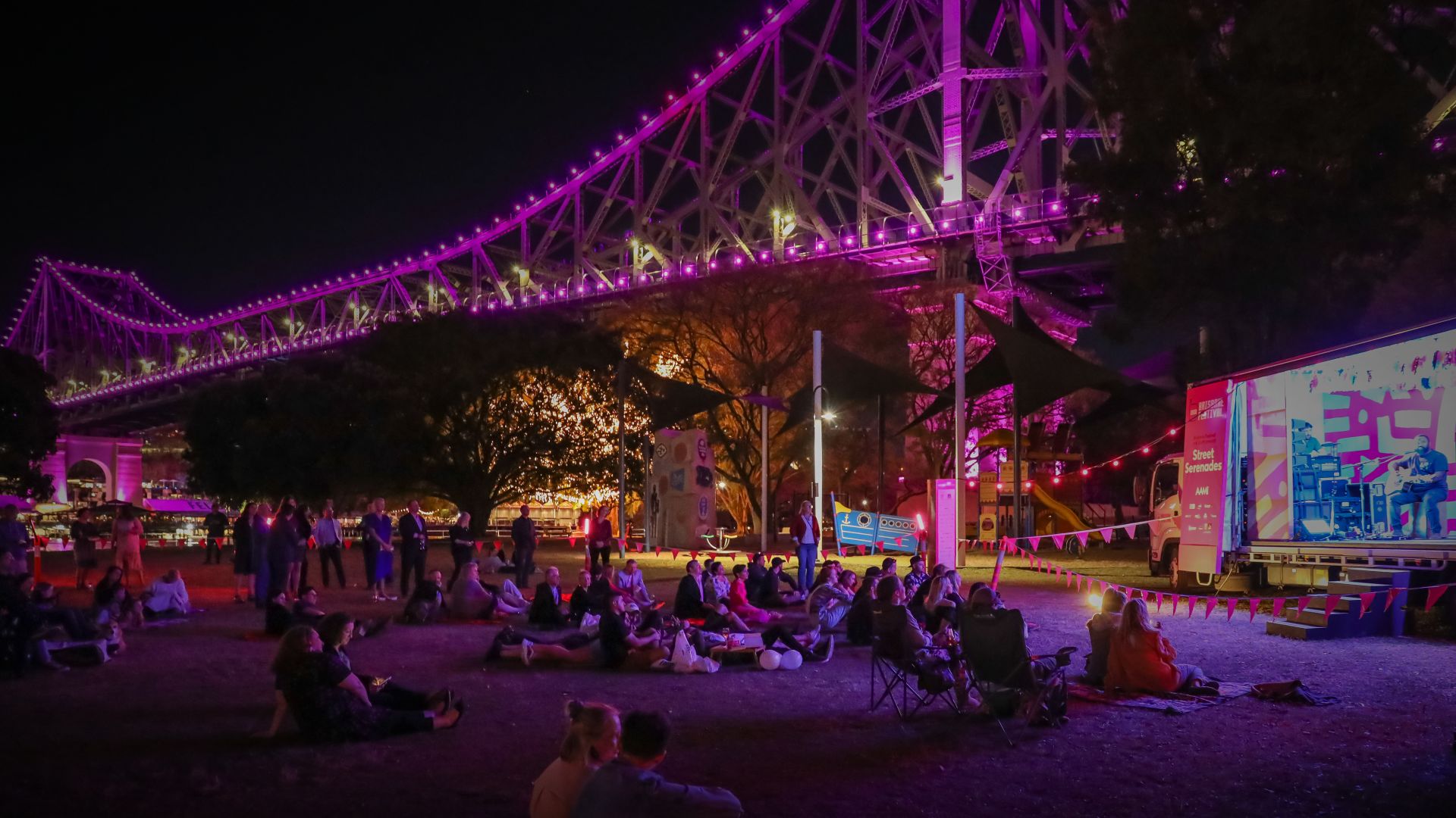 A socially distanced crowd watching musicians Busby Marou perform in a park by the Story Bridge at night time. 